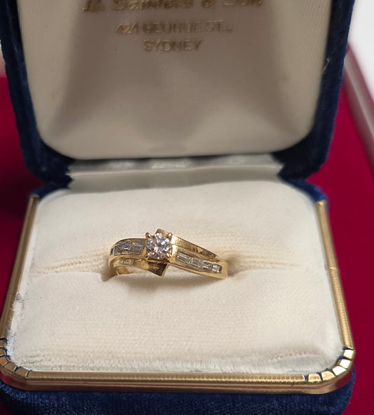18 ct Yellow gold bypass-style diamond ring featuring a round brilliant-cut center stone flanked by channel-set baguette or rectangular accent diamonds along the Size 7