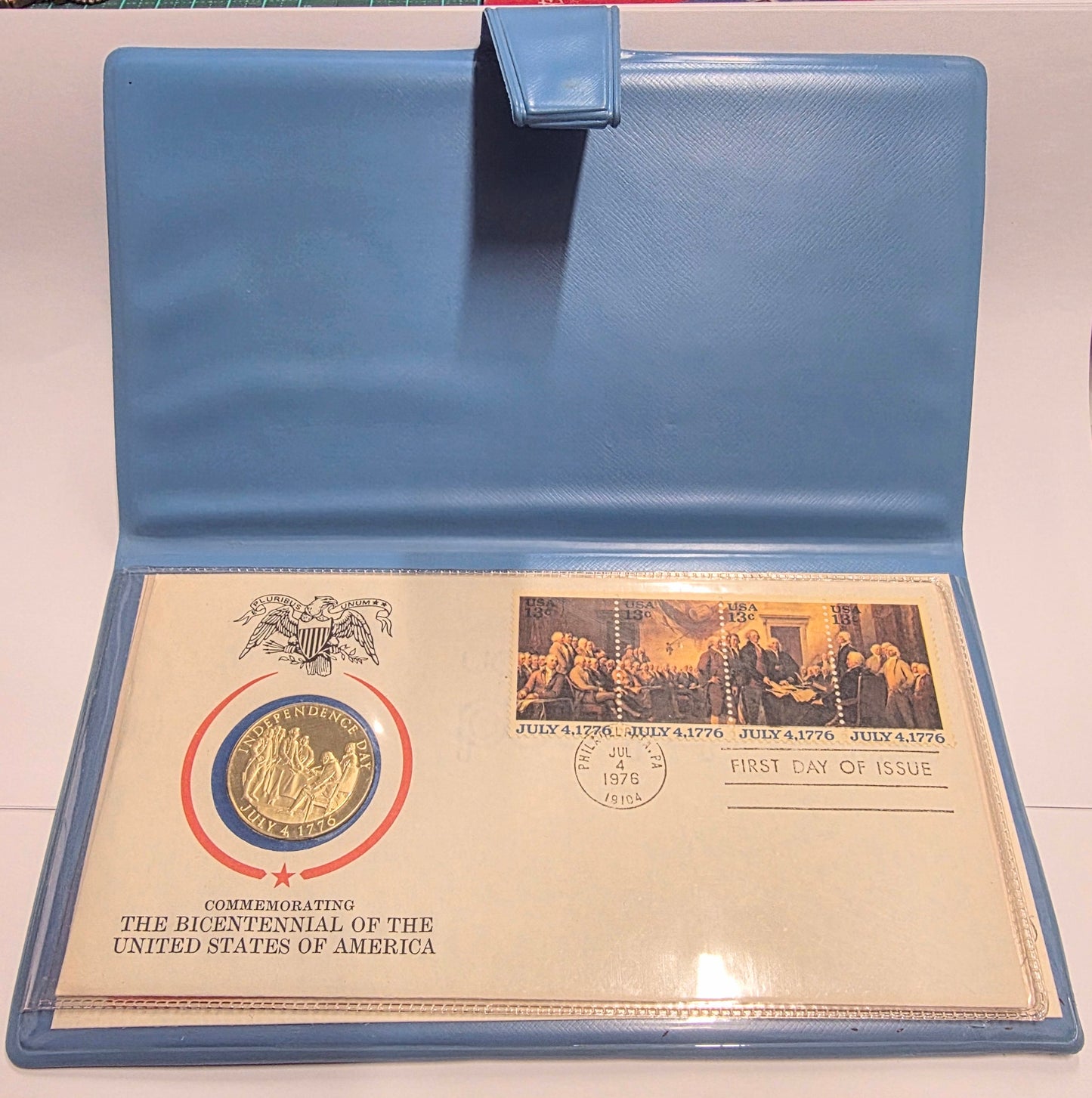 A light blue plastic document holder or envelope case shown open, containing a commemorative United States postage cover dated 1976. The cover includes a circular blue and red emblem, a black-and-white historical scene stamp, and printed text reading 'The Bicentennial of the United States of America' and 'First Day of Issue.